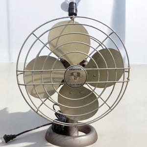 Puede incluir: Un ventilador vintage Emerson Electric con una bombilla en la parte superior. El ventilador tiene una jaula de metal y tres aspas. El ventilador es de color plateado.
