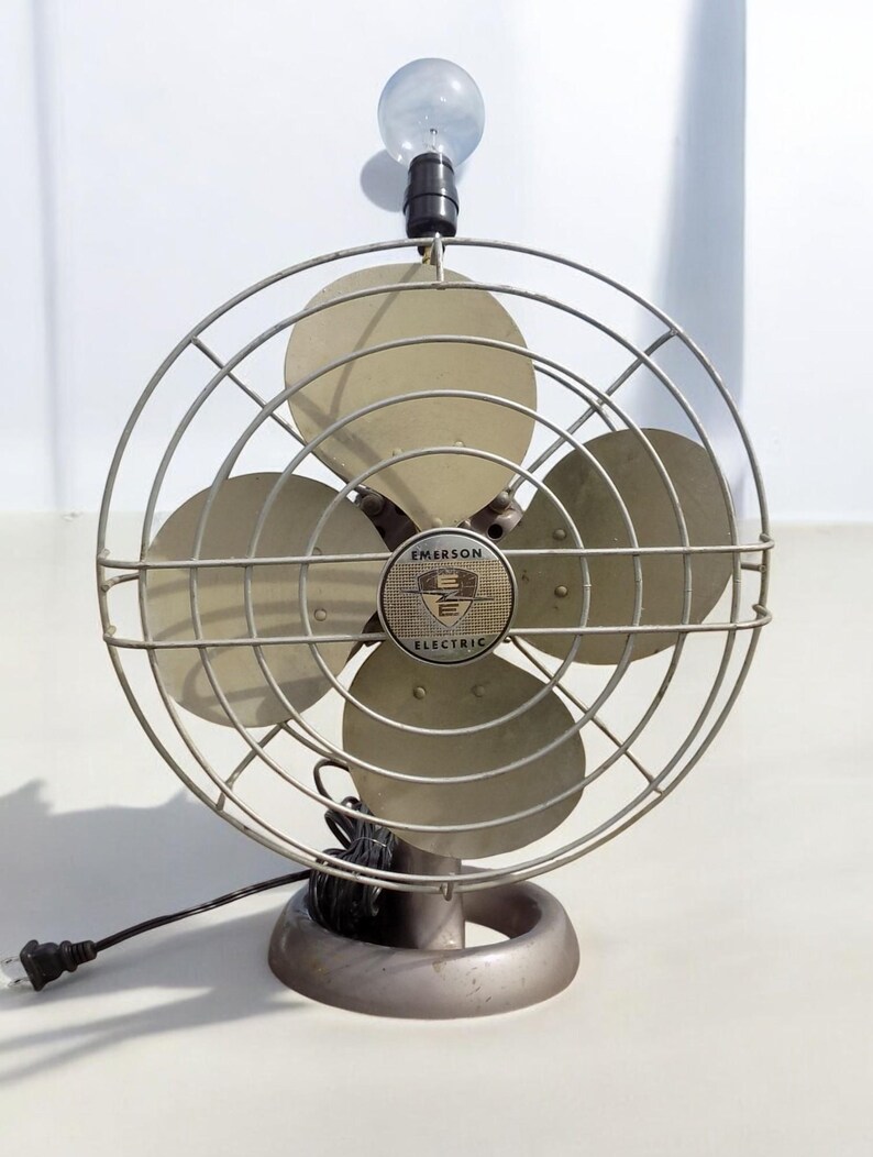 Puede incluir: Un ventilador vintage Emerson Electric con una bombilla en la parte superior. El ventilador tiene una jaula de metal y tres aspas. El ventilador es de color plateado.