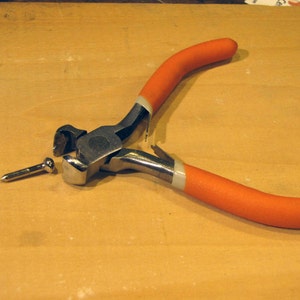Könnte beinhalten: Zange mit orangefarbenem Griff und silbernem Metallkopf, zum Schneiden von Draht oder Metall. Die Zange ist geöffnet und ein kleiner silberner Nagel liegt daneben.