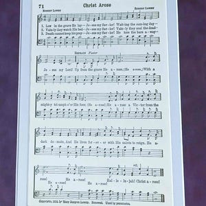 May include: A framed print of sheet music titled "Christ Arose." The music is printed in black ink on aged, cream-colored paper. The text includes lyrics and musical notation, with the composer's name at the top. The frame is a light blue color.