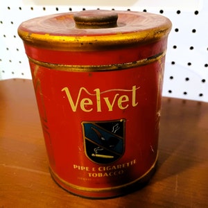 May include: Red metal tobacco tin with a gold lid and trim. The tin has a gold and white label with the word "Velvet" in a cursive font. The label also features a blue and white crest with a pipe and cigarette. The text "Pipe & Cigarette Tobacco" is printed below the crest.