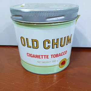 Puede incluir: Una lata de plata con una etiqueta verde que dice "Old Chum Cigarette Tobacco" y "Net Weight 200g".