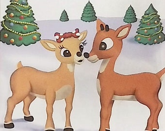 May include: A brown and white reindeer with a red bow on its head looks at a brown and white reindeer with a red nose. Both reindeer are standing in a snowy landscape with Christmas trees in the background.