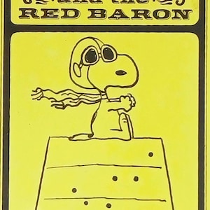 May include: Yellow graphic featuring Snoopy as the Red Baron, with the text "Snoopy and the Red Baron" in a black border. Snoopy is wearing goggles and a scarf, sitting on his doghouse.