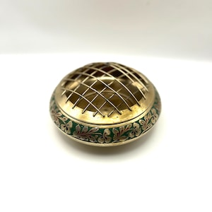 May include: A brass flower frog with a grid top and a green and floral patterned band. The flower frog is round and has a vintage aesthetic. The brass has a golden hue.