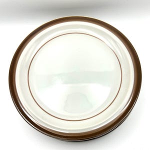 May include: A stack of white ceramic plates with a brown border and a thin brown accent line. The plates are round and appear to be stacked on top of each other. The plates are on a white background.