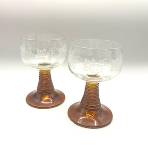 May include: Two clear wine glasses with etched grape and leaf designs. The stems and bases are amber-colored. The stems have a ribbed texture. The glasses are on a white background.