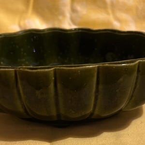 May include: A dark green ceramic bowl with a scalloped rim and vertical ribbed sides. The bowl has a glossy finish and sits on a yellow surface. This decorative bowl is suitable for serving or display.