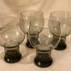May include: Four gray-tinted glass goblets with rounded bowls and short, dark bases. The glasses are arranged in a group, showcasing their simple, elegant design. The goblets are suitable for serving beverages.