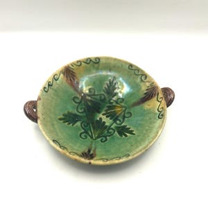 May include: A small, round, ceramic bowl with a green interior and a decorative floral design. The bowl has two small, brown handles and a scalloped edge. The bowl's exterior is a mix of green, brown, and yellow.