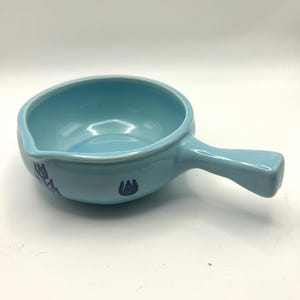 May include: A light blue ceramic sauce bowl with a handle. The bowl has a rounded shape with a pouring spout and is decorated with dark blue floral designs. The handle is rectangular and extends from the side of the bowl.