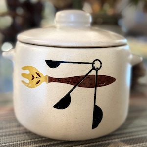 May include: Cream-colored ceramic cookie jar with a lid and two side handles. The jar features a retro design with a yellow fork, brown spoon, and black measuring spoons. The speckled surface adds a vintage feel.