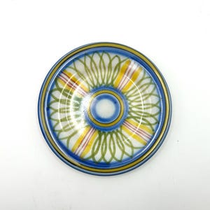 May include: A round, decorative ceramic plate with a colorful design. The plate features a blue rim and a central pattern of concentric circles and lines in yellow, green, and red. The plate is approximately 6 inches in diameter.
