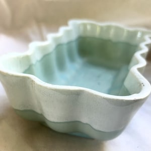 May include: A ceramic dish with a wavy, scalloped edge. The dish is painted in shades of light blue and white, with a contrasting darker blue interior. The handmade pottery has a rustic, artisanal feel.