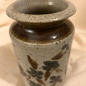 May include: A ceramic vase with a speckled, light gray body and a wide, rounded rim. The vase features a dark brown band near the top and is decorated with blue and brown floral patterns. The vase has a cylindrical shape.