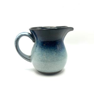 May include: A handmade ceramic pitcher with a rounded body and a handle. The pitcher features a gradient of colors, transitioning from a dark blue rim to a light blue-green base. The spout is designed for easy pouring.