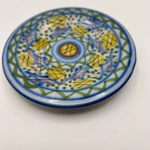 May include: A small, round ceramic plate with a hand-painted design. The plate features a blue rim and a central pattern of yellow, green, and red accents. The design includes geometric shapes and floral elements, creating a decorative piece.