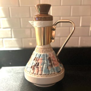 May include: A vintage ceramic pitcher with a brown, gold, and white design. The pitcher has a handle and a spout. The lower portion of the pitcher features a mosaic pattern in blue, pink, and brown. The lid is brown.
