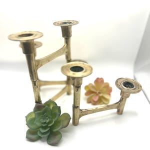 May include: A vintage brass candle holder with four candle cups. The candle holder has an angular design with a gold finish. A green succulent and a small pink flower are in the foreground.