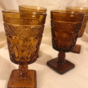 May include: Set of four amber-colored vintage water goblets. Each glass features a square base, a textured stem, and a detailed diamond and floral pattern on the bowl. The glasses are made of thick glass.