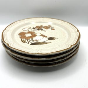 May include: A stack of five ceramic plates with a floral design. The plates have a scalloped edge and a speckled beige background. The floral pattern features brown and white flowers and leaves.