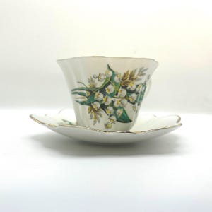 May include: A white porcelain teacup and saucer set featuring a scalloped edge and a gold-tone rim. The teacup is decorated with a botanical illustration of green lily of the valley leaves and small white bell-shaped flowers on the exterior.