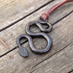 May include: A black metal snake pendant necklace with a brown leather cord. The pendant is a simple, stylized design of a snake with a rounded head.