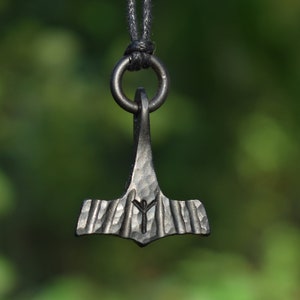 May include: A dark metal pendant in the shape of a hammer, suspended from a black cord. The hammer head features etched lines and a rune symbol. The background is a blurred green, suggesting an outdoor setting. The pendant is likely a piece of jewelry.