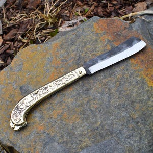 Peut inclure: Un couteau pliant avec un manche en laiton orné d'un motif floral complexe. La lame est en acier et présente une légère courbure.