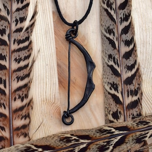 May include: Black metal bow pendant necklace on a black cord. The pendant is a simple, curved shape with a small ring at the bottom. The necklace is displayed on a wooden surface with pheasant feathers.