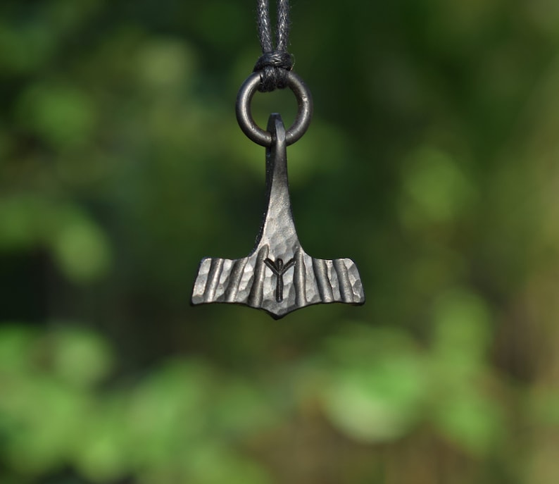 May include: A dark metal pendant in the shape of a hammer, suspended from a black cord. The hammer head features etched lines and a rune symbol. The background is a blurred green, suggesting an outdoor setting. The pendant is likely a piece of jewelry.
