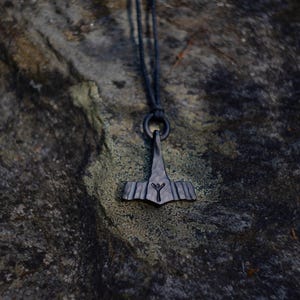 May include: A dark gray pendant necklace featuring a stylized hammer design, suspended from a black cord. The pendant has a textured surface and a rune symbol. The necklace is displayed against a mottled, dark stone background.