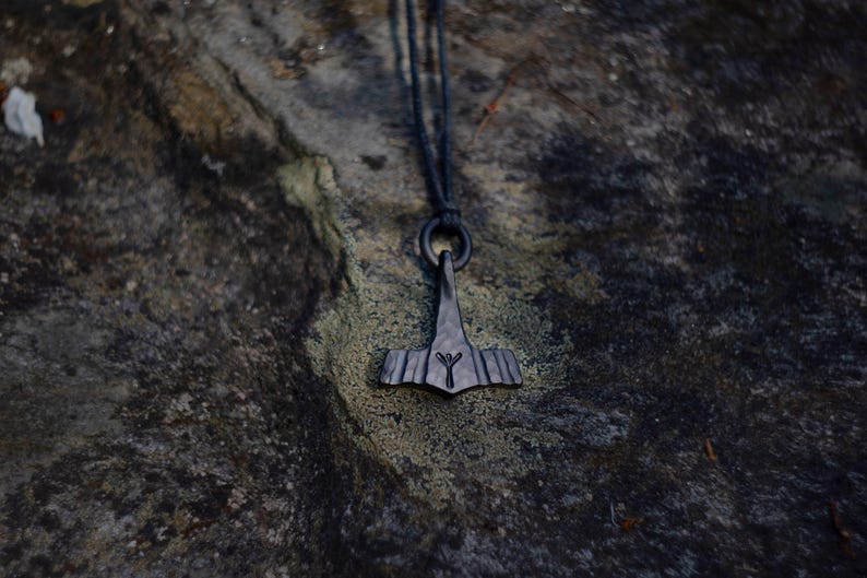 May include: A dark gray pendant necklace featuring a stylized hammer design, suspended from a black cord. The pendant has a textured surface and a rune symbol. The necklace is displayed against a mottled, dark stone background.