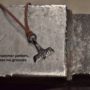 May include: A dark metal pendant in the shape of Thor's hammer, with a textured surface, hangs from a brown leather cord. The pendant is suspended against a textured metal surface. Text on the surface reads: "Plain hammer pattern, no runes no grooves."