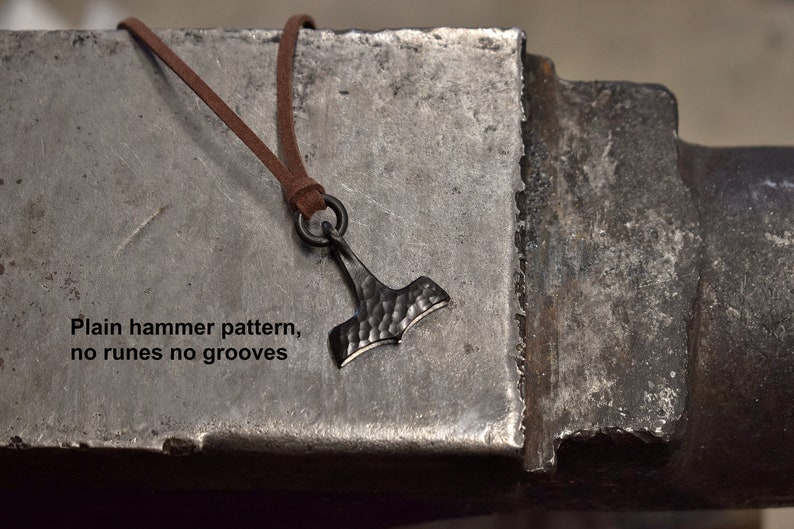 May include: A dark metal pendant in the shape of Thor's hammer, with a textured surface, hangs from a brown leather cord. The pendant is suspended against a textured metal surface. Text on the surface reads: "Plain hammer pattern, no runes no grooves."