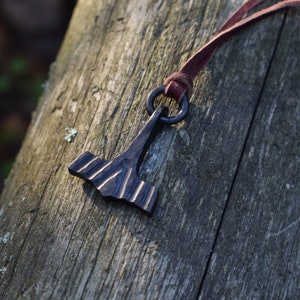 May include: A dark metal pendant in the shape of a hammer, with a textured surface and a dark brown leather cord. The pendant is suspended from a metal ring. The hammer is resting on a weathered wooden surface.