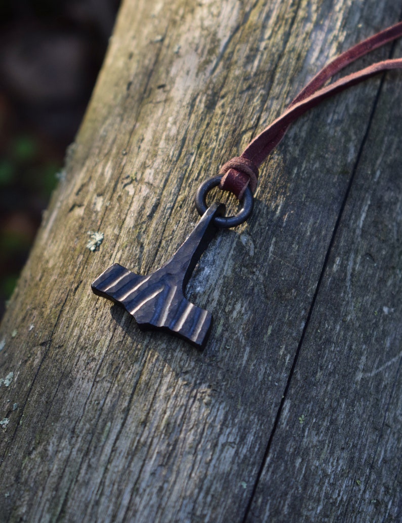 May include: A dark metal pendant in the shape of a hammer, with a textured surface and a dark brown leather cord. The pendant is suspended from a metal ring. The hammer is resting on a weathered wooden surface.
