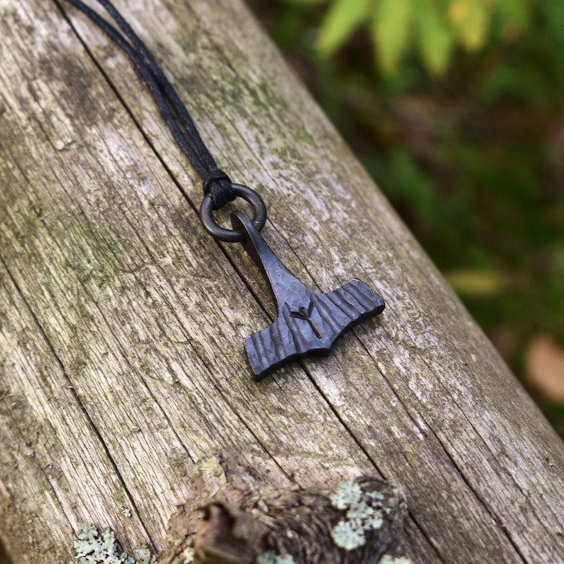 May include: A black metal pendant necklace featuring a hammer design, with a rune symbol etched into the head. The pendant hangs from a black cord, and the background is a weathered wooden log.