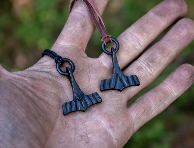 May include: Two black metal Thor's hammer pendants. Each pendant is suspended from a black cord or brown leather strap. The hammer design features a textured head and a simple handle. The background is blurred, suggesting an outdoor setting.
