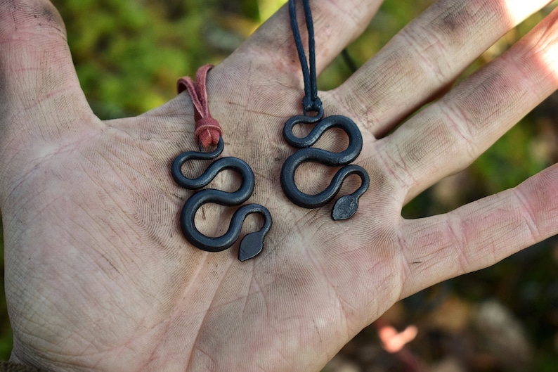 May include: Two black snake pendant necklaces on a hand. One necklace has a red cord and the other has a black cord.