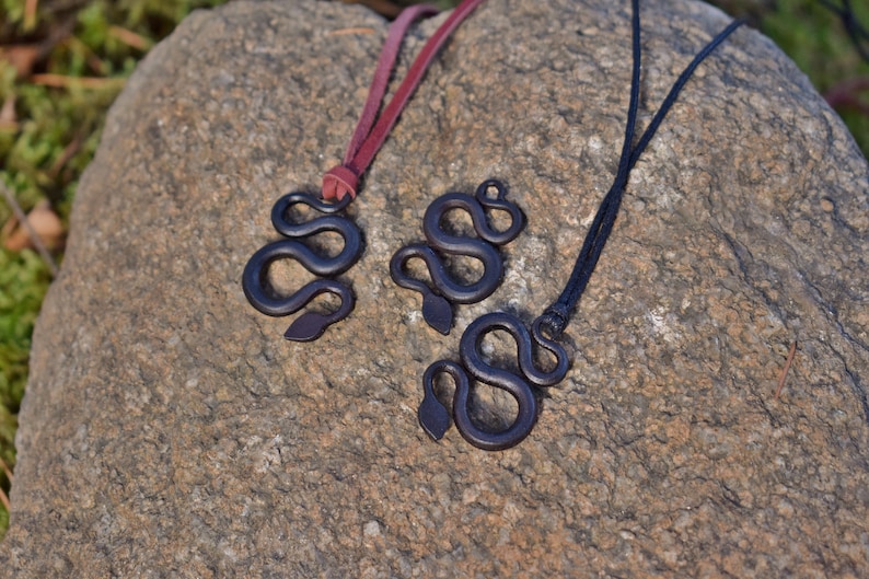 May include: Three black snake pendant necklaces on a rock background. Two of the necklaces have brown leather cords and one has a black cord.
