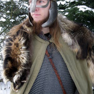 May include: A medieval-style outfit featuring a silver helmet with a face guard, chainmail, a green cloak, and a fur collar. A brown leather belt and strap are also visible. The background shows evergreen trees and snow.