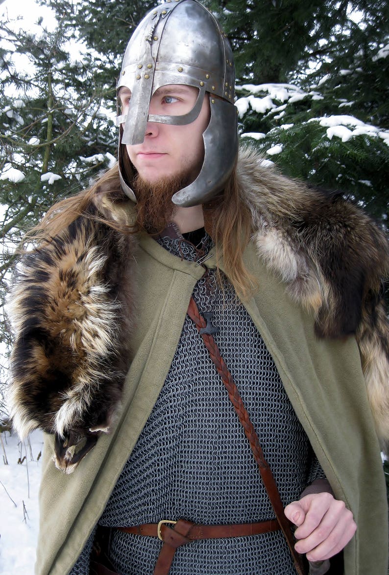 May include: A medieval-style outfit featuring a silver helmet with a face guard, chainmail, a green cloak, and a fur collar. A brown leather belt and strap are also visible. The background shows evergreen trees and snow.