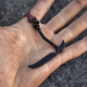 May include: A black metal hook with a curved blade, attached to a brown leather strap. The hook is held in a person's hand.