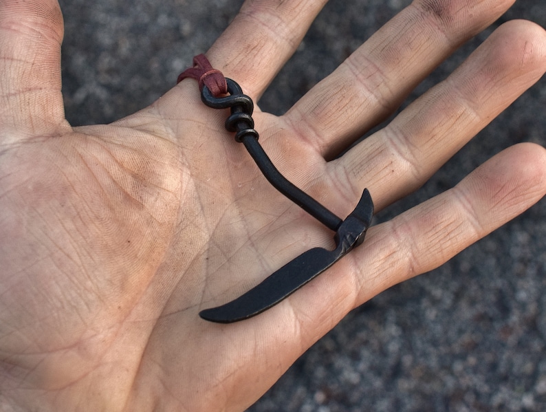 May include: A black metal hook with a curved blade, attached to a brown leather strap. The hook is held in a person's hand.
