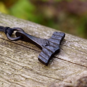 May include: A dark gray, forged metal pendant in the shape of Thor's hammer, Mjolnir, hangs from a black cord. The hammer has a textured surface and a small symbol etched into the head. The pendant rests on a weathered wooden surface.