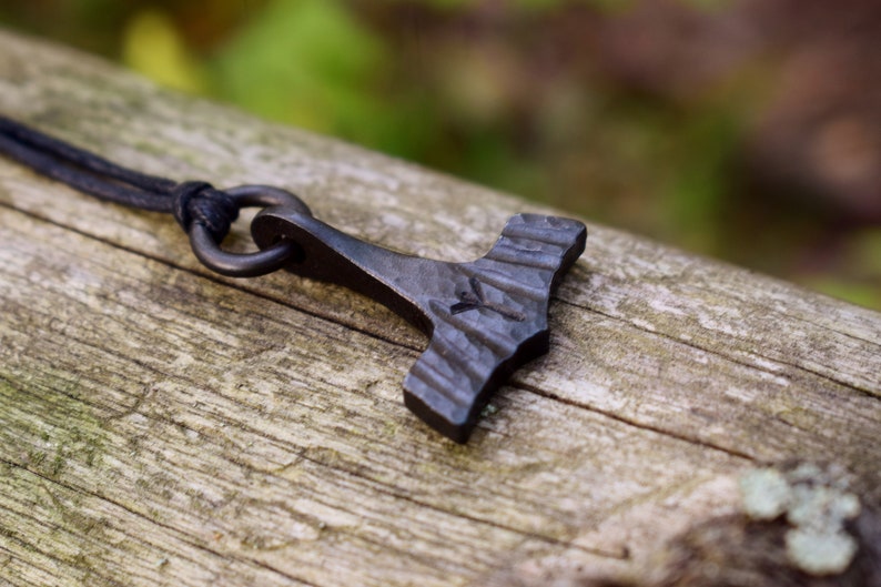 May include: A dark gray, forged metal pendant in the shape of Thor's hammer, Mjolnir, hangs from a black cord. The hammer has a textured surface and a small symbol etched into the head. The pendant rests on a weathered wooden surface.