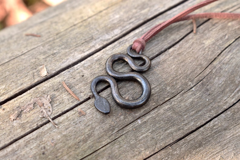 May include: A black metal snake pendant necklace with a brown leather cord. The pendant is a simple, stylized design of a snake with a rounded head.
