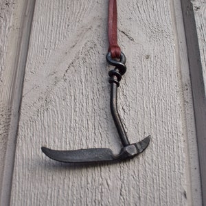 May include: A black metal hook pendant with a brown leather cord. The hook is shaped like a curved blade with a pointed tip.