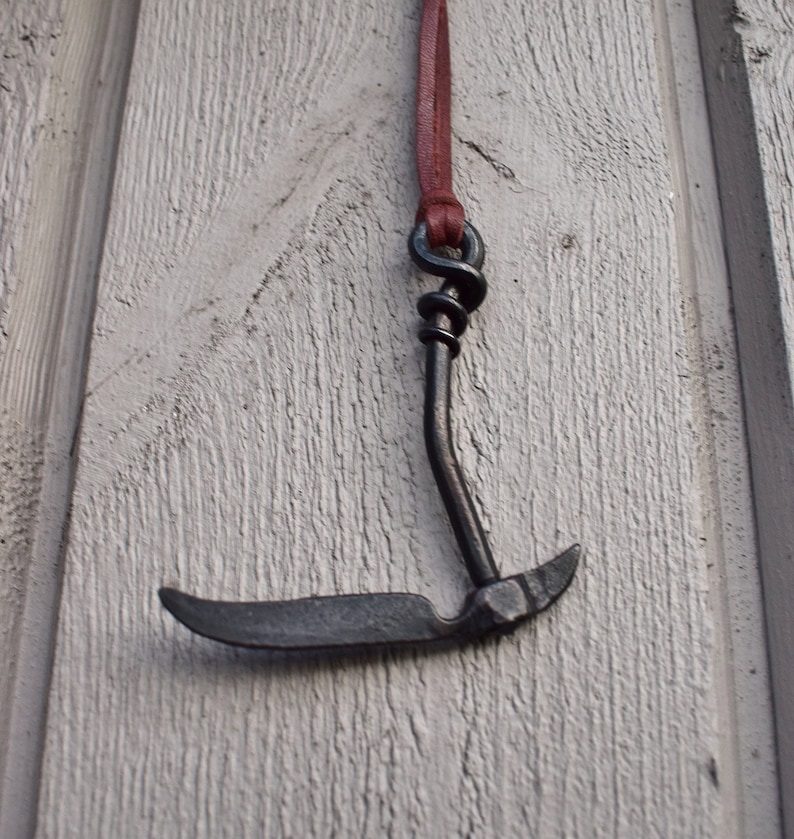 May include: A black metal hook pendant with a brown leather cord. The hook is shaped like a curved blade with a pointed tip.